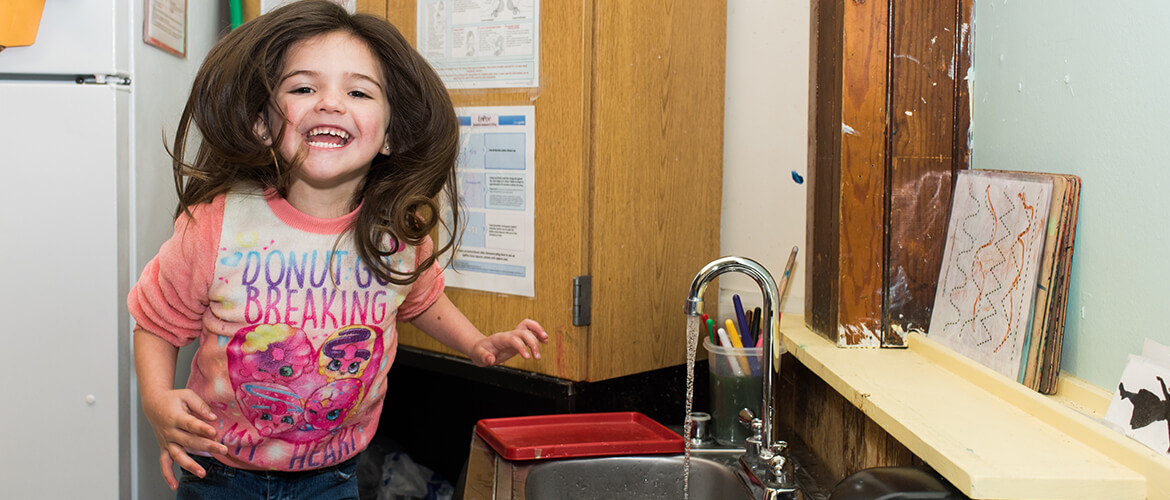 Young child smiling next to a sink with running water