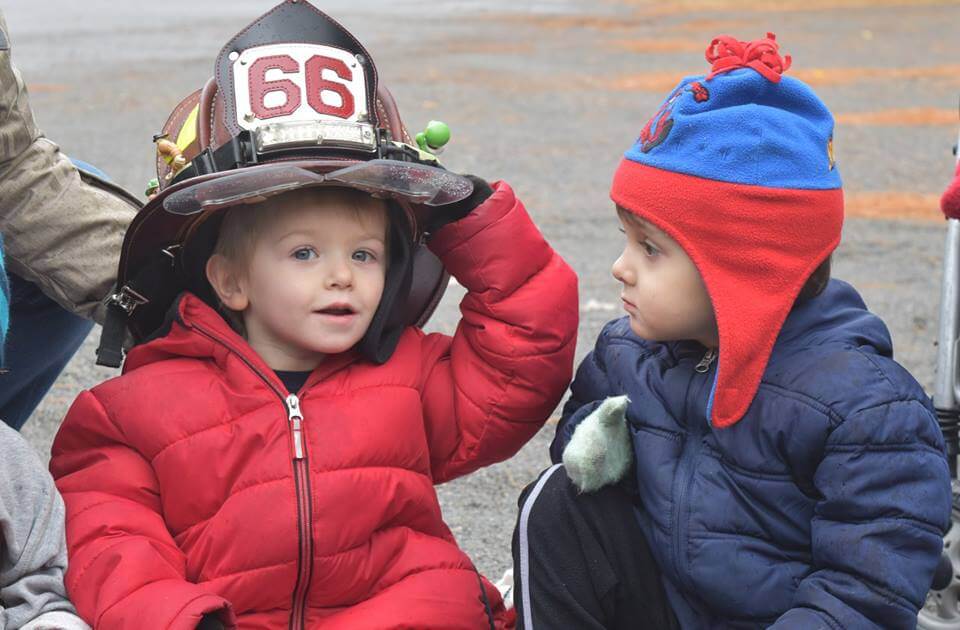 A little boy in a red coat smiles as he tries on a real firefighter's hat