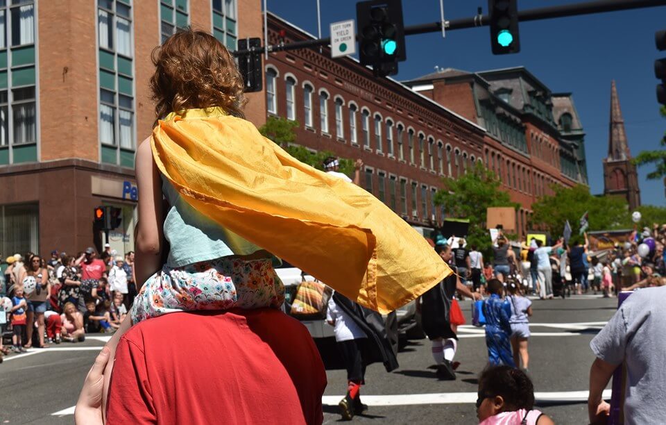 View from behind of a little girl perched atop a man's shoulders in the parade. The little girl wears a yellow cape that is fluttering in the wind.